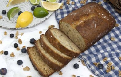 loaf-of-sliced-banana-bread-garnished-with-blueberries-powdered-sugar-walnuts-and-citrus-on-checkered-tablecloth