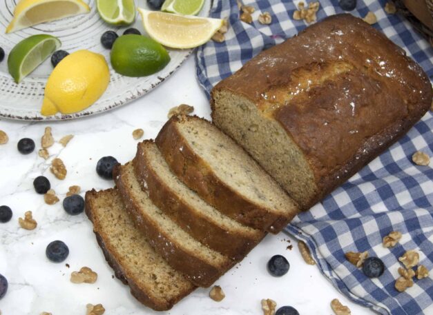 loaf-of-sliced-banana-bread-garnished-with-blueberries-powdered-sugar-walnuts-and-citrus-on-checkered-tablecloth